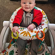 Dakota joined the competition — help win amazing prizes! baby, infant, smiling, baby_rocker, toy_rings, colorful, red_clothing, gray_clothing, carpet, indoor, happy, child, person, seat, pattern, leaf_pattern, mushroom_pattern, hand, footwear, playtime