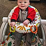 baby, infant, smiling, baby_rocker, toy_rings, colorful, red_clothing, gray_clothing, carpet, indoor, happy, child, person, seat, pattern, leaf_pattern, mushroom_pattern, hand, footwear, playtime