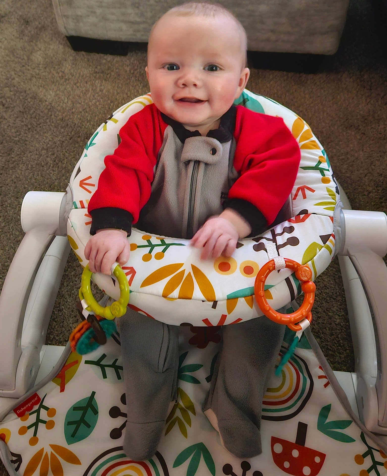 Dakota joined the competition — help win amazing prizes! baby, infant, smiling, baby_rocker, toy_rings, colorful, red_clothing, gray_clothing, carpet, indoor, happy, child, person, seat, pattern, leaf_pattern, mushroom_pattern, hand, footwear, playtime