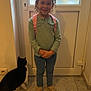 child, smiling, curly_hair, indoor, door, frosted_glass, backpack, pink, green_shirt, blue_pants, shoes, tile_floor, cat, black_and_white_cat, pet, happy, standing, curious, home, person