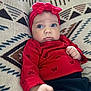baby, blue_eyes, bow, child, closeup, cozy, curious_expression, cute, face, fist, headband, indoors, infant, patterned_cushion, portrait, red_clothing, sitting, soft_lighting, textile_pattern, young