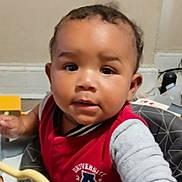 Ezekiel is registered to the contest to win money with this photo: baby, child, infant, portrait, face, indoors, high_chair, seated, red_clothing, gray_sleeves, curious, serious_expression, looking_at_camera, short_hair, dark_skin, household, furniture, toy, close_up, photography