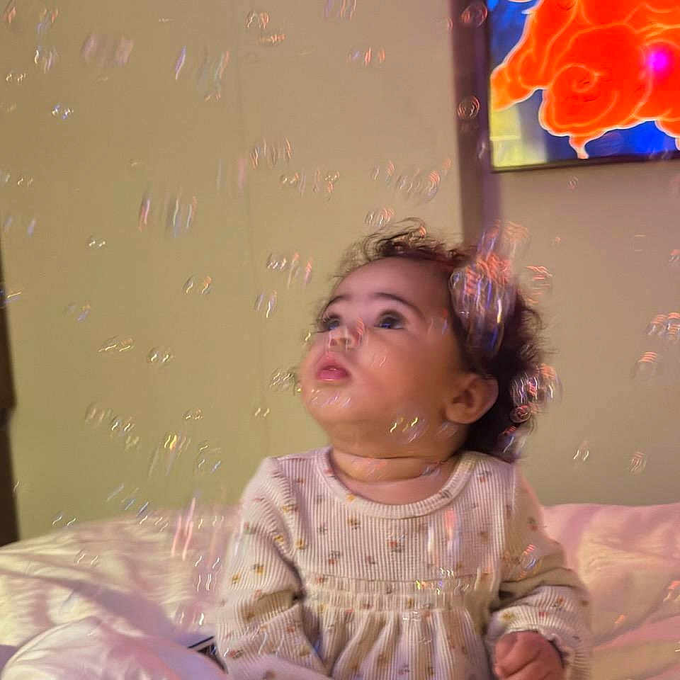Asha joined the competition — help win amazing prizes! baby, bed, bubbles, child, clothing, colorful, curious, cute, gazing, indoor, infant, long_sleeve, painting, portrait, room, sitting, small_hands, soft_lighting, wall, young_child