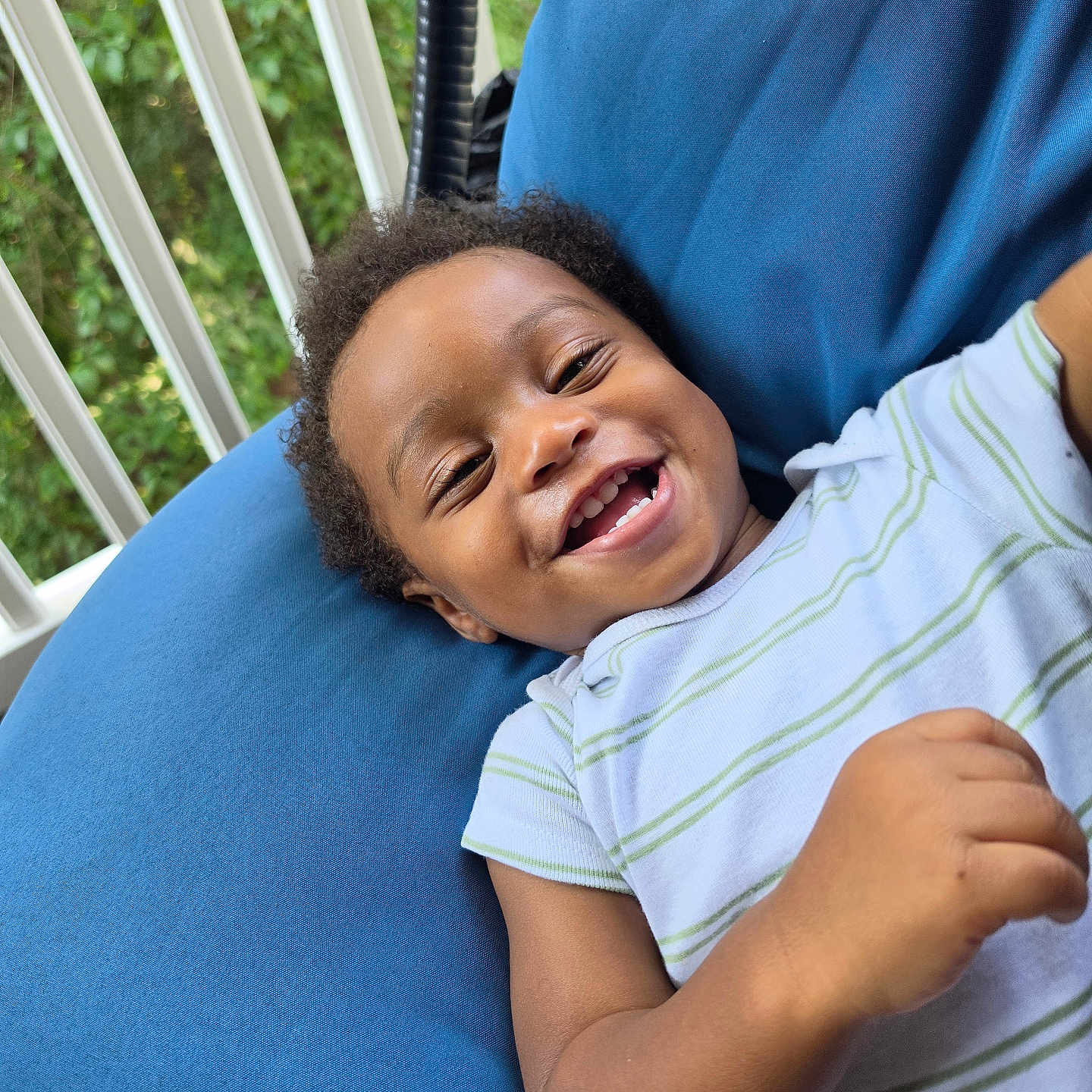 Elijah is registered to the contest to win money with this photo: baby, child, smiling, laughing, portrait, face, teeth, curly_hair, sitting, lying_down, blue_cushion, striped_shirt, short_sleeves, outdoor, balcony, railing, greenery, daylight, happy, close_up