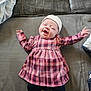 baby, infant, newborn, smiling, face, hat, plaid_dress, dress, socks, leggings, arms_up, lying_down, couch, blanket, fabric, home_interior, portrait, happy, soft_lighting, cute