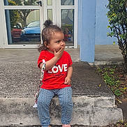 Maelys a rejoint le concours — aidez-le/la à gagner de superbes lots ! casual_clothing, child, concrete_step, curiosity, curly_hair, daylight, glass_door, hand_on_mouth, love_text, outdoor, patterned_pants, pavement, pink_sandals, plants, red_shirt, reflection, side_view, toddler, tree, urban