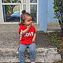 casual_clothing, child, concrete_step, curiosity, curly_hair, daylight, glass_door, hand_on_mouth, love_text, outdoor, patterned_pants, pavement, pink_sandals, plants, red_shirt, reflection, side_view, toddler, tree, urban