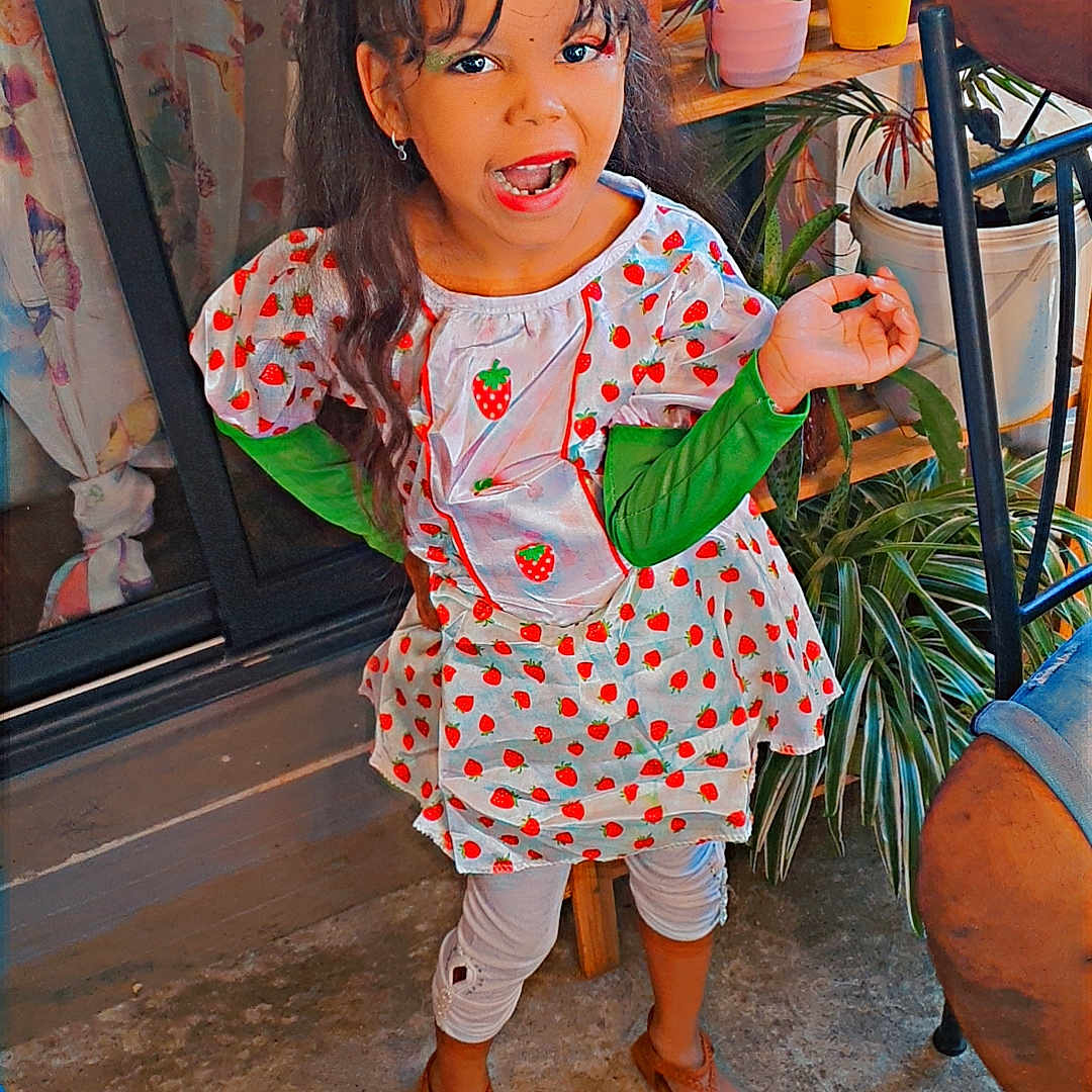 Luna a rejoint le concours — aidez-le/la à gagner de superbes lots ! child, concrete_floor, curtain, dress, earrings, expression, footwear, girl, green_sleeves, hair, home, indoor, long_hair, person, plants, potted_plants, sandals, shelf, smiling, strawberry_pattern