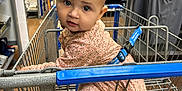Ariyah is registered to the contest to win money with this photo: baby, shopping_cart, clothing, store, aisle, child, infant, curious, face, person, retail, floor, metal, blue, pink, patterned_clothing, safety_belt, head, eyes, expression