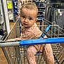 baby, shopping_cart, clothing, store, aisle, child, infant, curious, face, person, retail, floor, metal, blue, pink, patterned_clothing, safety_belt, head, eyes, expression