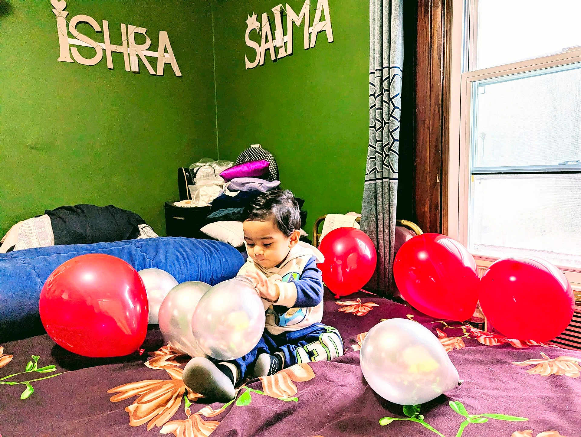 Shadab is registered to the contest to win money with this photo: child, toddler, balloon, bed, blanket, window, curtain, green_wall, pillow, clothing, floor, indoor, furniture, person, room, light, toy, celebration, sitting, play