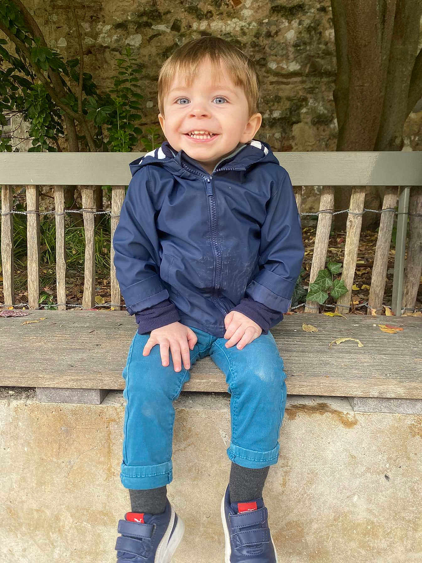 Malo participe au concours pour gagner de l'argent avec cette photo : child, toddler, smiling, blue_eyes, jacket, pants, shoes, bench, outdoor, nature, tree, leaves, stone_wall, happy, person, casual_clothing, sitting, footwear, daylight, cute