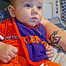 arm, baby, bedroom, bib, child, cute, drool, expression, face, hand, indoor, infant, orange_clothing, person, photostrip, portrait, purple_bib, sitting, tattoo, wall