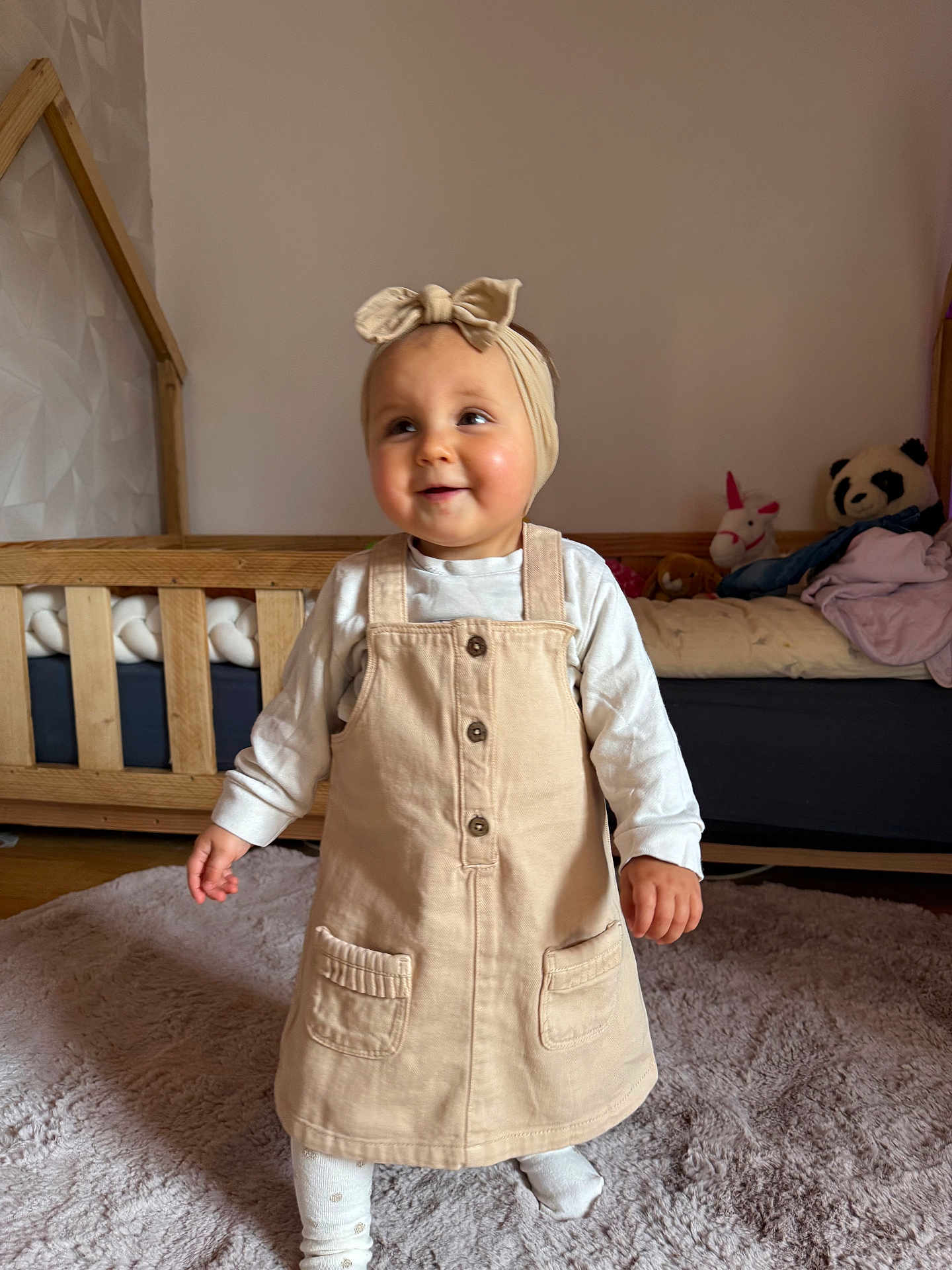 Gwendoline participe au concours pour gagner de l'argent avec cette photo : toddler, child, beige_dress, headband, carpet, wooden_crib, stuffed_animals, panda_toy, unicorn_toy, indoor, smiling, happy, curious, baby, person, clothing, floor, room, long_sleeve_shirt, footwear