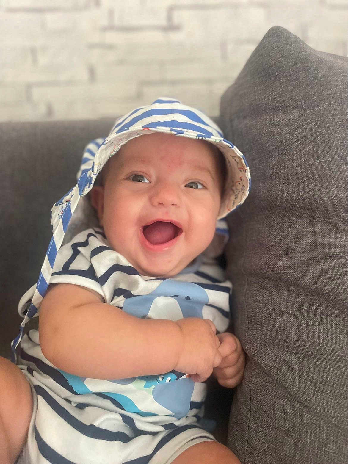 Noé participe au concours pour gagner de l'argent avec cette photo : baby, child, smiling, happy, hat, striped_clothing, couch, indoor, infant, cute, portrait, person, sitting, young_child, joyful, face, expression, casual, comfortable, relaxed