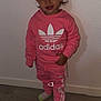 toddler, child, pink_clothing, adidas, sweatshirt, pants, socks, standing, smiling, indoor, wooden_floor, wall, hair, face, person, casual, cute, fashion, young_child, portrait