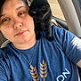 person, kitten, black_cat, car_interior, selfie, blue_shirt, necklace, cross_pendant, affection, pet, cute, animal, closeup, indoor, seatbelt, window, daylight, snuggling, face, shoulder