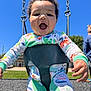 Kingston is registered to the contest to win money with this photo: baby, child, swing, playground, smiling, curly_hair, outdoor, blue_sky, sunny, clothing, dinosaur_print, person, happy, joyful, daylight, play, fun, park, seat, infant