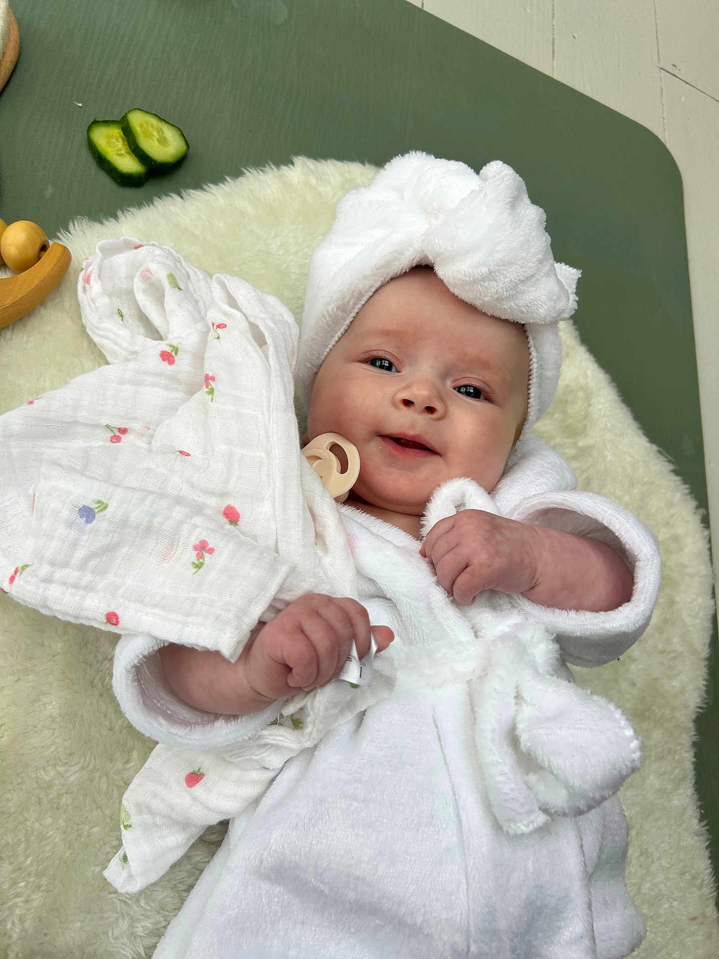 Marliah is registered to the contest to win money with this photo: baby, smiling, white_robe, towel_turban, blanket, cucumber_slices, hand, face, infant, soft_texture, cozy, lying_down, cute, skin, indoors, comfort, person, headwear, pacifier, portrait