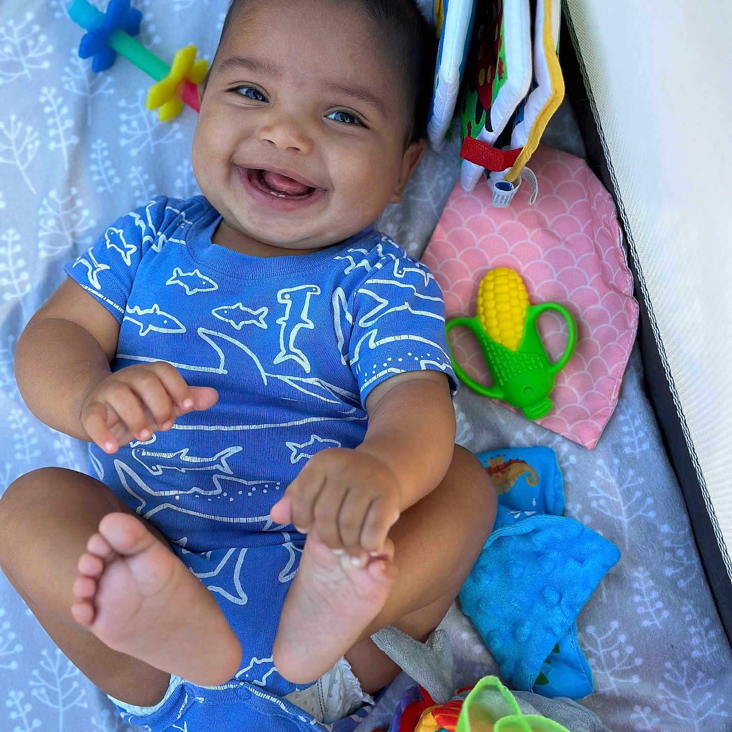 Ezekiel joined the competition — help win amazing prizes! baby, blanket, blue_onesie, child, colorful, cute, face, feet, hand, happy, indoor, infant, lying_down, playtime, portrait, shark_pattern, smiling, soft_toys, teether, toys