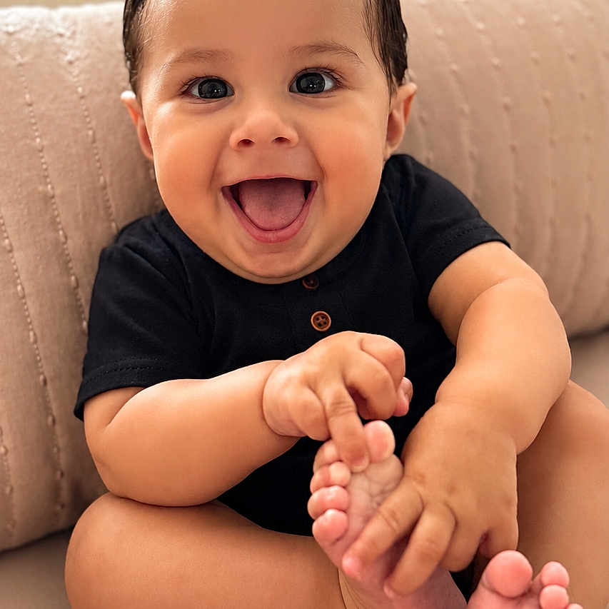 Jimmy is registered to the contest to win money with this photo: baby, child, smiling, happy, sitting, foot, hand, couch, pillow, indoor, cute, person, infant, black_clothing, skin, expression, portrait, young, toes, playful