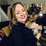 woman, dog, puppy, smiling, portrait, indoor, living_room, person, happy, black_clothing, bracelet, earrings, cute, pet, home_decor, sofa, cushion, blonde_hair, holding, face