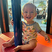 Kayden is registered to the contest to win money with this photo: baby, toddler, smile, happy, play_area, playground, mesh_screen, blue_pole, red_seat, onesie, barefeet, sitting, sunlight, portrait, child, joyful, cute, teeth, indoor_playground, grid_background