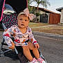 toddler, stroller, child, outdoor, sidewalk, street, headband, pink_bow, cartoon_clothing, sneakers, person, tree, car, house, gate, fence, palm_tree, curious, casual, daytime