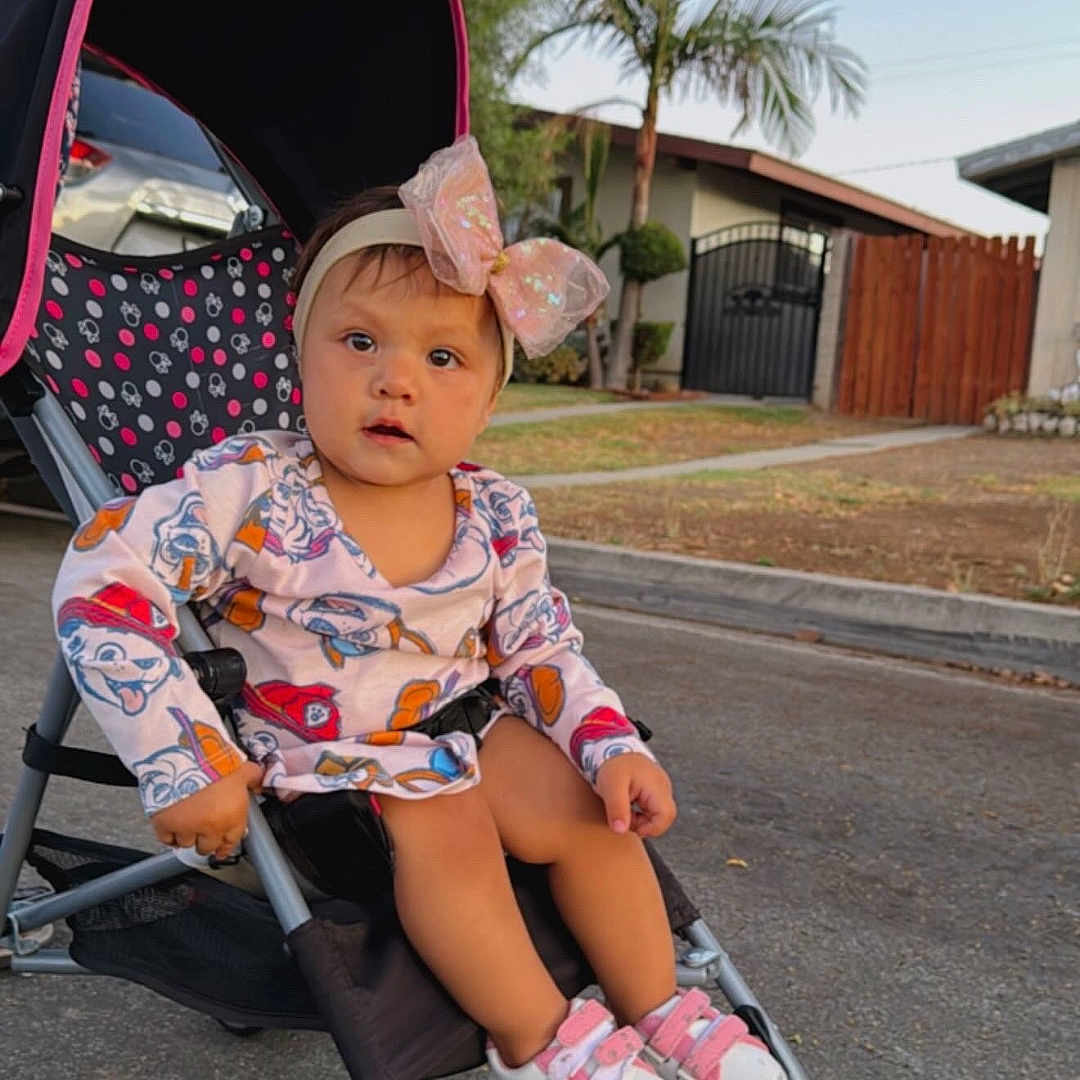 Zaraiht Daynela joined the competition — help win amazing prizes! car, cartoon_clothing, casual, child, curious, daytime, fence, gate, headband, house, outdoor, palm_tree, person, pink_bow, sidewalk, sneakers, street, stroller, toddler, tree
