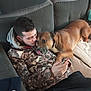affection, brown_dog, camouflage_jacket, casual, comfort, couch, cozy, dog, friendship, furniture, indoor, interaction, leisure, man, pet, phone, playing, relaxing, seated, young_adult
