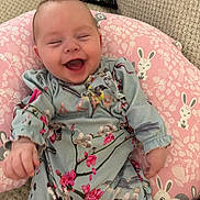Mabel joined the competition — help win amazing prizes! baby, laughing, smiling, infant, pink_pillow, bunny_pattern, floral_clothing, blue_outfit, knitted_blanket, remote_control, happy, cute, child, indoor, portrait, face, person, sitting, cozy, soft