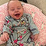baby, laughing, smiling, infant, pink_pillow, bunny_pattern, floral_clothing, blue_outfit, knitted_blanket, remote_control, happy, cute, child, indoor, portrait, face, person, sitting, cozy, soft