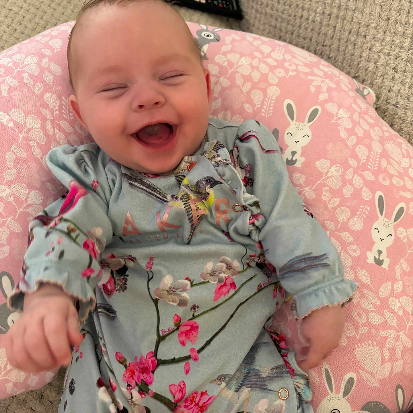 Mabel joined the competition — help win amazing prizes! baby, blue_outfit, bunny_pattern, child, cozy, cute, face, floral_clothing, happy, indoor, infant, knitted_blanket, laughing, person, pink_pillow, portrait, remote_control, sitting, smiling, soft
