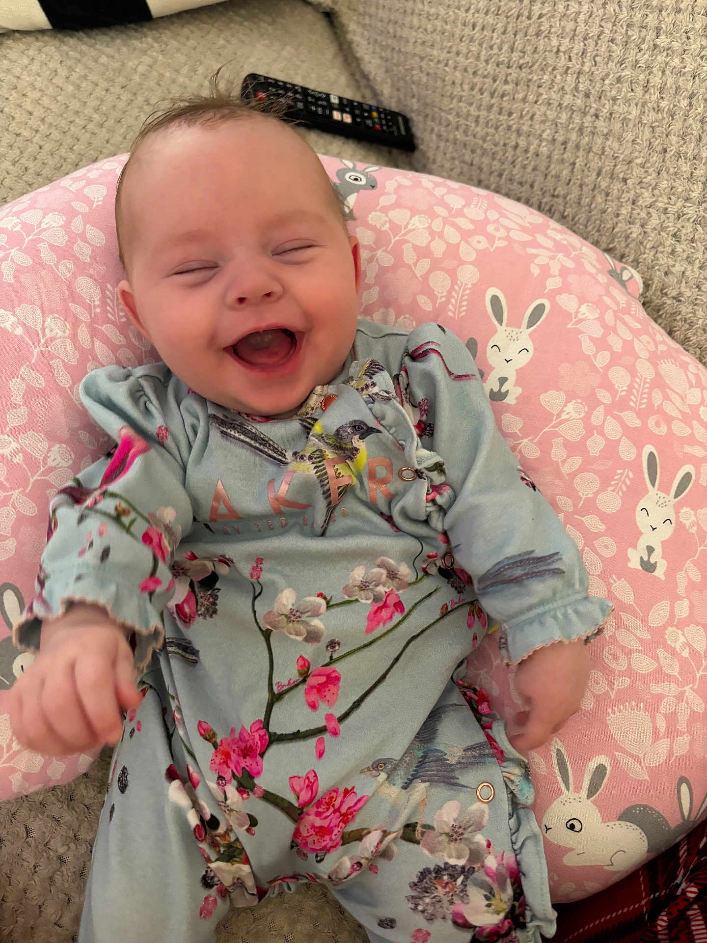 Mabel joined the competition — help win amazing prizes! baby, laughing, smiling, infant, pink_pillow, bunny_pattern, floral_clothing, blue_outfit, knitted_blanket, remote_control, happy, cute, child, indoor, portrait, face, person, sitting, cozy, soft