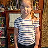 child, girl, portrait, standing, smiling, striped_shirt, ponytail, hair_bow, indoors, home, cabinet, furniture, figurines, decorations, mirror, curtain, carpet, collection, front_facing, photograph