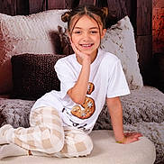 Saisha is registered to the contest to win money with this photo: child, girl, smile, portrait, indoor, pajamas, plaid_pants, white_shirt, hair_buns, bracelet, cozy, ottoman, pillow, wood_background, cushion, happy, teeth, sitting, studio_shot, face