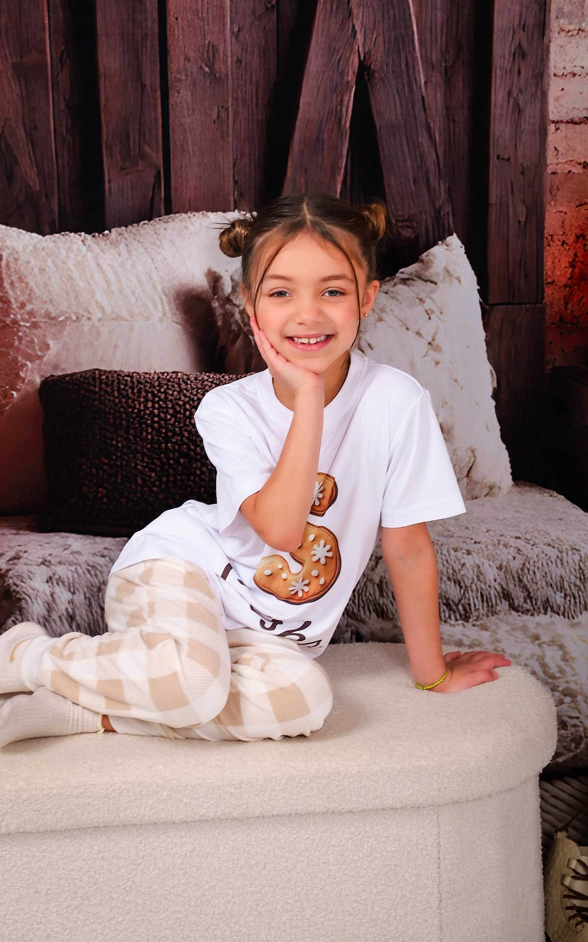 Saisha is registered to the contest to win money with this photo: child, girl, smile, portrait, indoor, pajamas, plaid_pants, white_shirt, hair_buns, bracelet, cozy, ottoman, pillow, wood_background, cushion, happy, teeth, sitting, studio_shot, face