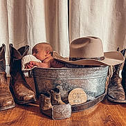 Wesson is registered to the contest to win money with this photo: baby, newborn, metal_tub, cowboy_boots, cowboy_hat, wooden_floor, curtains, milestone, one_month, belt, wooden_plaque, infant, portrait, indoors, rustic, decor, flooring, clothing, hat, footwear