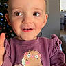 toddler, child, smiling, purple_sweatshirt, bear_graphic, flower, indoor, christmas_tree, holiday_decor, face, eyes, expression, cute, happy, portrait, young_child, sitting, home, cozy, long_sleeves