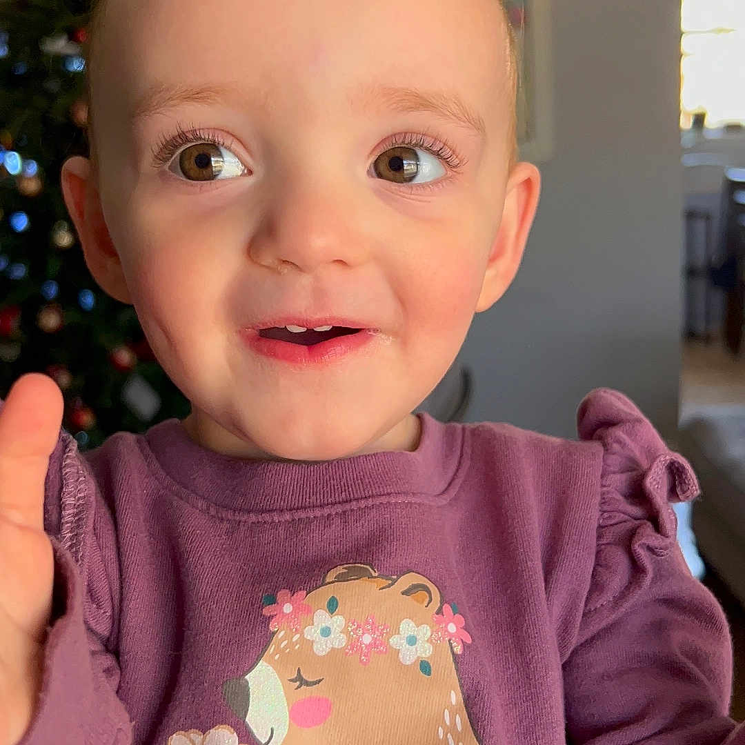 Emma Ruth joined the competition — help win amazing prizes! toddler, child, smiling, purple_sweatshirt, bear_graphic, flower, indoor, christmas_tree, holiday_decor, face, eyes, expression, cute, happy, portrait, young_child, sitting, home, cozy, long_sleeves