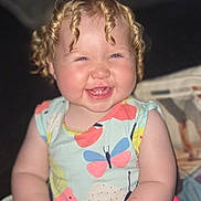 Hallie joined the competition — help win amazing prizes! baby, child, smiling, curly_hair, blonde, dress, butterfly_pattern, sitting, happy, indoor, couch, person, infant, cute, hands, legs, portrait, young_child, skin, expression