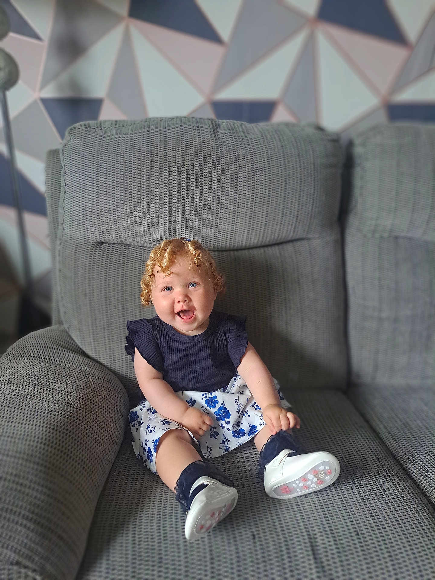 Hallie joined the competition — help win amazing prizes! toddler, child, smiling, curly_hair, sitting, couch, gray_couch, blue_top, floral_skirt, white_shoes, indoor, happy, portrait, person, furniture, home, patterned_wall, geometric_pattern, cute, baby