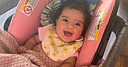 Olivia is registered to the contest to win money with this photo: baby, bib, blanket, car_seat, child, comfort, couch, cozy, cute, face, happy, indoor, infant, living_room, person, pink_blanket, seated, smiling, soft_texture, warm_light