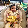 Olivia is registered to the contest to win money with this photo: baby, child, smile, curly_hair, yellow_skirt, white_top, baby_chair, cushion, indoor, blue_couch, wooden_floor, green_baby_seat, furniture, happy, person, footwear, sock, home, play_area, cute