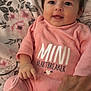 baby, infant, pink_onesie, smiling, face, hand, tattoo, blanket, floral_pattern, cute, child, lying_down, indoor, person, clothing, skin, portrait, happy, young_child, cozy