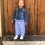Léna a rejoint le concours — aidez-le/la à gagner de superbes lots ! child, smile, denim_jacket, pants, shoes, stone_step, wooden_door, standing, outdoor, happy, person, fashion, casual, portrait, cute, red_hair, clothing, footwear, background, wall