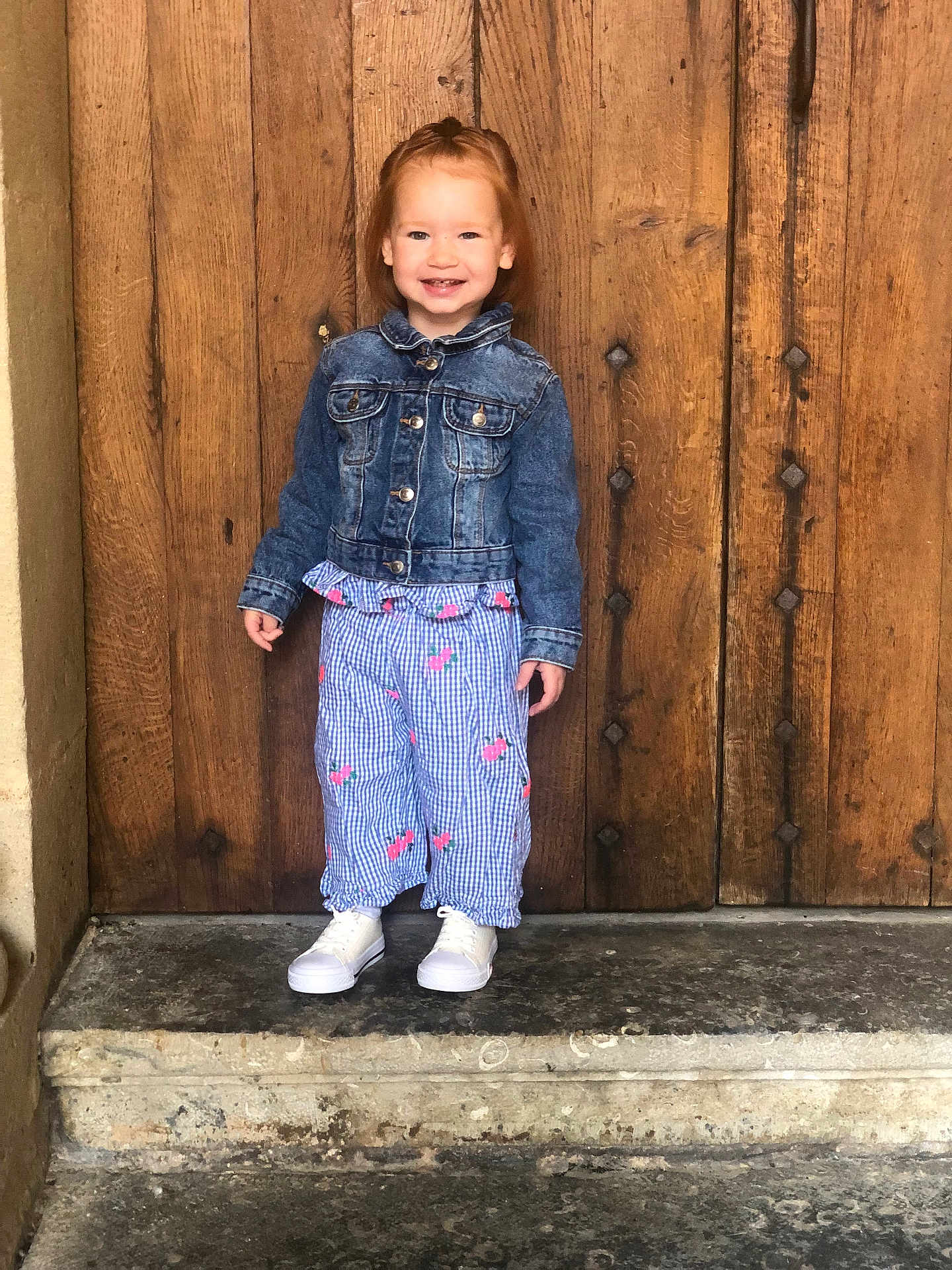 Léna a rejoint le concours — aidez-le/la à gagner de superbes lots ! child, smile, denim_jacket, pants, shoes, stone_step, wooden_door, standing, outdoor, happy, person, fashion, casual, portrait, cute, red_hair, clothing, footwear, background, wall