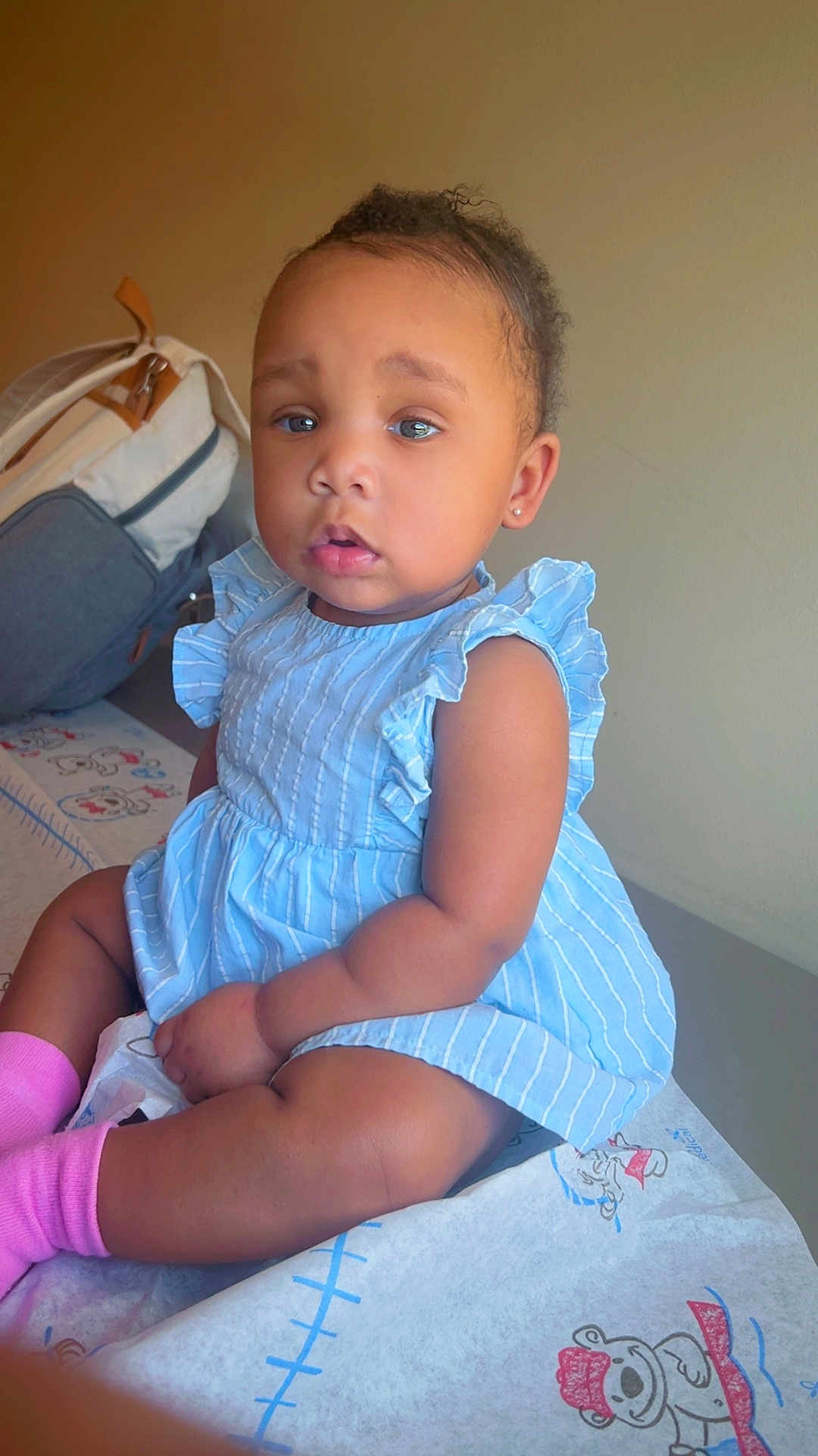 Autumn is registered to the contest to win money with this photo: toddler, child, baby, blue_dress, pink_socks, sitting, indoor, curly_hair, earring, teddy_bear_print, sheet, light, expression, cute, person, portrait, young_child, skin, face, clothing