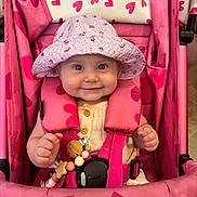 Nola is registered to the contest to win money with this photo: baby, child, smiling, stroller, pink, hat, toy, heart_pattern, safety_harness, cute, infant, clothing, seat, happy, person, portrait, indoor, beads, wooden_toy, fabric