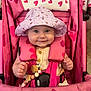 baby, child, smiling, stroller, pink, hat, toy, heart_pattern, safety_harness, cute, infant, clothing, seat, happy, person, portrait, indoor, beads, wooden_toy, fabric